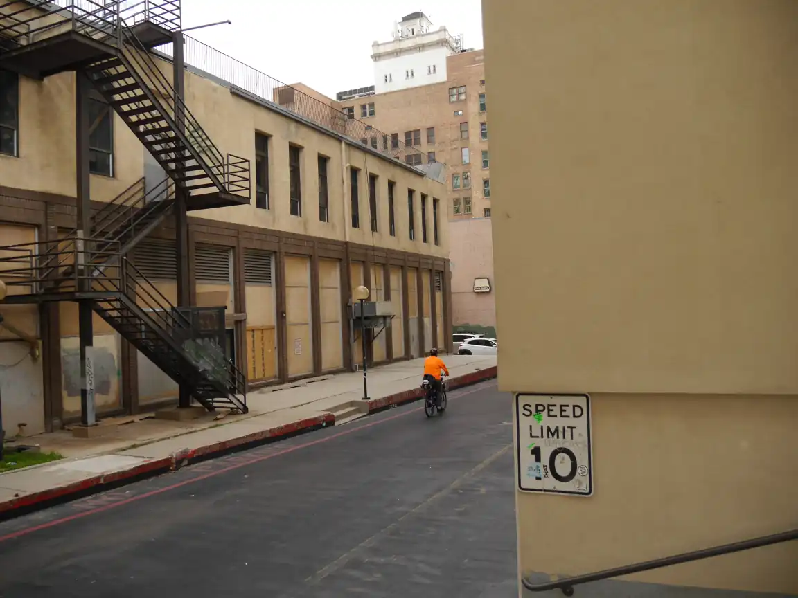 A person in a flourescent orange jacket bicycling out of an alleyway, framed by a 'SPEED LIMIT 10' sign